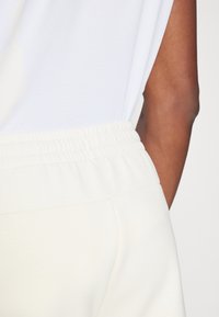 Vit elastisk midja med samlad textur och mjukt tyg; ett par ljuskrämfärgade byxor med slät yta, som har sidofickor.