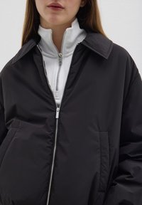 Schwarze gepolsterte Jacke mit breitem Kragen, vorderem Reißverschluss und Seitentasche. Darüber wird ein weißer Fleece-Reißverschluss mit hohem Kragen getragen.