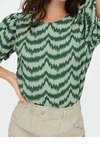 Blouse verte à motifs avec des rayures en zigzag, manches courtes bouffantes et coupe décontractée, associée à un pantalon beige taille haute.
