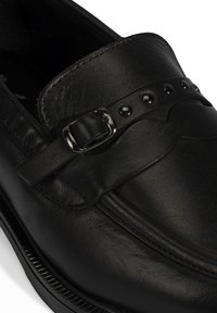 Zwarte leren loafers met een gestructureerd oppervlak, een decoratieve riem met een gesp, en ronde zwarte studs langs de bovenrand.