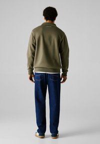 Sudadera verde oliva, corte relajado, puños y dobladillo acanalados. Combinada con jeans oscuros de corte suelto. Camisa blanca debajo, con un branding mínimo.