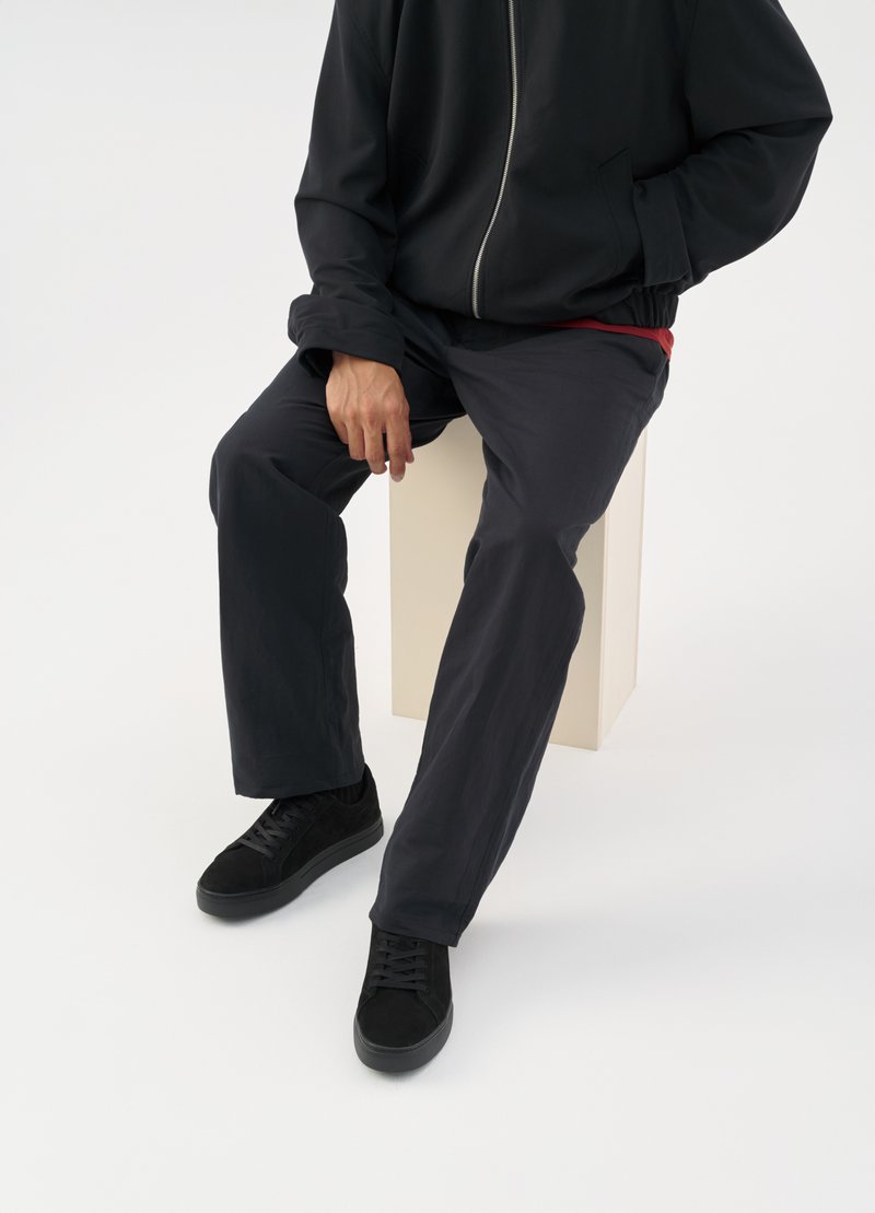 Μαύρο σακάκι με φερμουάρ, κόκκινο πουκάμισο ορατό από κάτω, σκούρο παντελόνι και μαύρα παπούτσια με κορδόνια. Άνδρας καθισμένος σε ένα φωτεινό βάθρο.