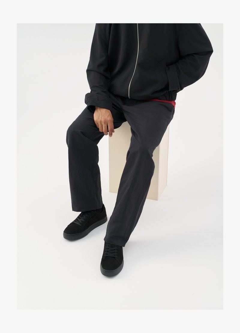 Fekete cipzáras dzseki, alatta vörös ing látható, sötét nadrág és fekete fűzős tornacipő. Férfi egy világos színű pedesztálon ül.