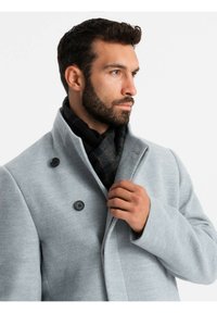 Cappotto di lana grigio con colletto alto, dotato di bottoni neri e design semi-strutturato, abbinato a una sciarpa a quadri scuri.