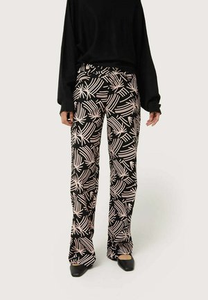 Pantalones negros con un patrón de hojas de palmera blancas, de forma acampanada, textura suave y detalle en la cinturilla, combinados con una blusa negra holgada y zapatos negros de punta.