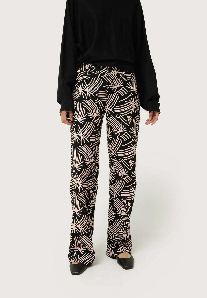 Pantalones negros con un patrón de hojas de palmera blancas, de forma acampanada, textura suave y detalle en la cinturilla, combinados con una blusa negra holgada y zapatos negros de punta.