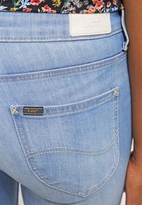 Jeans de denim azul claro con una etiqueta de cuero blanca que presenta "Lee". Un bolsillo trasero tiene un borde cosido y una pequeña etiqueta negra.