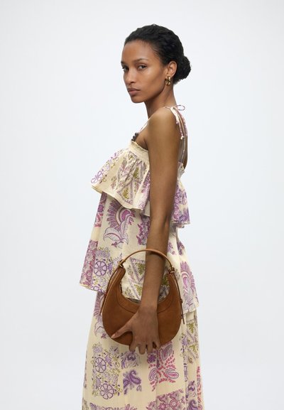 Femme en robe fleurie sans manches avec des volants, tenant un sac à main marron en forme de croissant, debout de profil sur un fond uni.