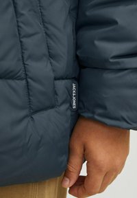 Zilā pufainā jaka ar izšūtu tekstūru, ar zīmola uzlīmi "JACK & JONES" uz piedurknes, uzvilkta virs bēša krekla.