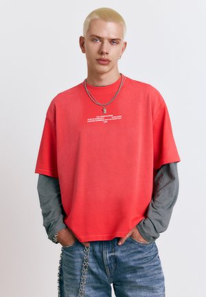 Ung person med kort blondt hår iført en rød oversized T-shirt over en langærmet, stribet skjorte og blå jeans med kæde-accessory.