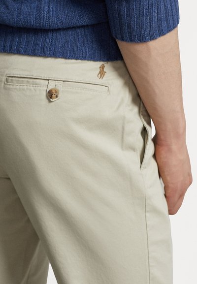 Beige bomullsbukser med en baklomme, som har en liten brun knapp og brodert hestelogo over lommen.