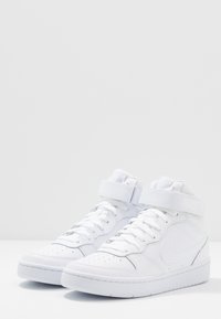 Nike högstövlar i helt vit läder. Funktioner inkluderar vadderad vrist, snörning fram, kardborreband och perforerad tåkapp.