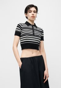 Νεαρό άτομο με κοντά σκούρα μαλλιά φοράει ριγέ μαύρο-άσπρο κοντό πόλο πουκάμισο και φαρδιά μαύρη παντελόνι, ποζάροντας μπροστά σε ένα απλό φόντο.