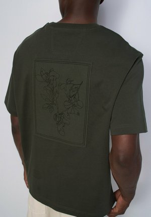 Camisa de manga corta de color verde oscuro que lleva una persona, con un diseño botánico cosido enmarcado en un rectángulo en la parte superior de la espalda.