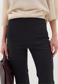 Zwarte aansluitende broek van rekbaar materiaal, gecombineerd met een crèmekleurige oversized trui. Een bruine handtas in de linkerhand.