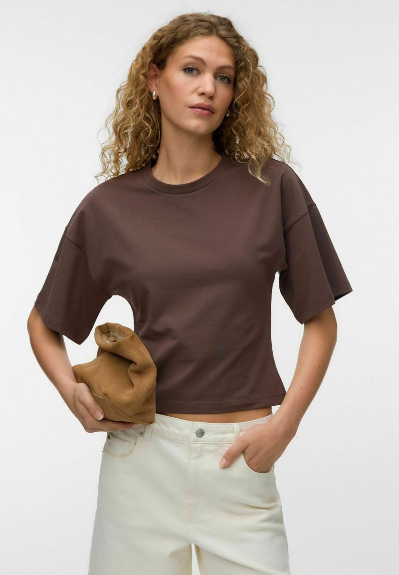Femme aux cheveux bouclés porte un t-shirt court ample marron et un pantalon blanc, tenant une pochette en daim beige pliée.