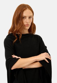 Czarny wełniany poncho z wysokim kołnierzem, luźnymi rękawami i swobodnym krojem. Teksturowany materiał o gładkim wykończeniu i minimalnych detalach.