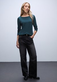 Teal langærmet top med bådudskæring, parret med højtaljede sorte brede jeans. Modellen står med den ene hånd i lommen.