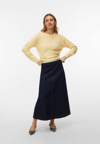 Jersey amarillo claro, falda larga de mezclilla oscura con detalles en las costuras y tacones puntiagudos con patrón. Presenta texturas de tela suave.