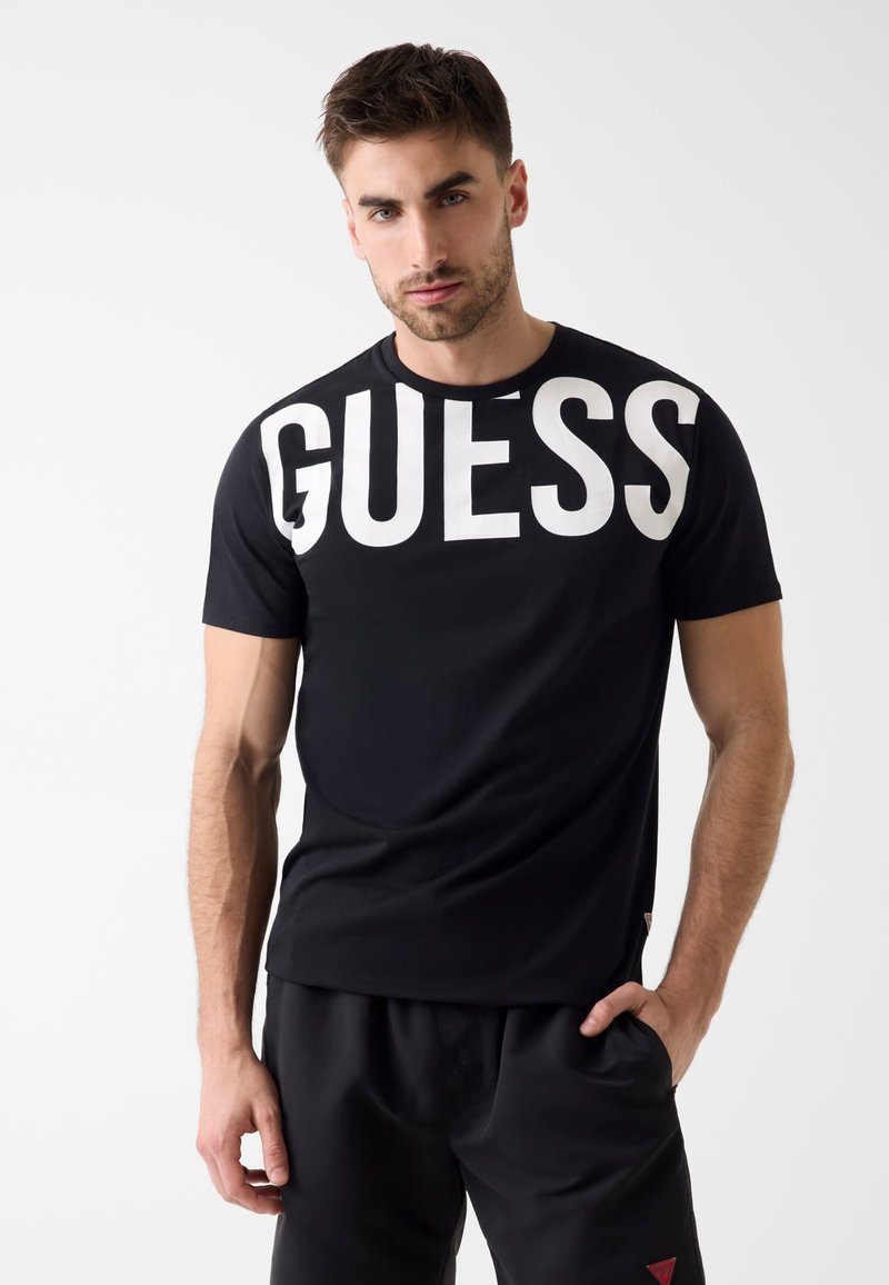 Uomo che indossa una maglietta nera con la grande scritta bianca "GUESS" sul petto e pantaloni neri, posando con una mano in tasca.