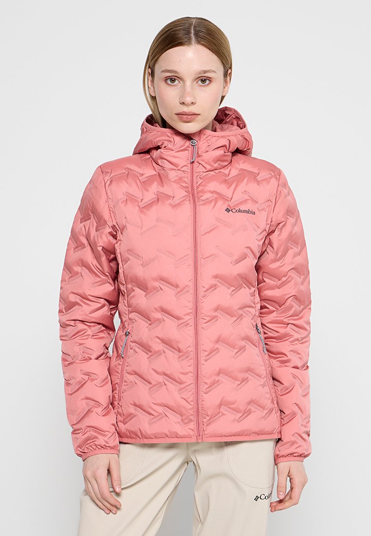 Mujer vestida con una chaqueta con capucha acolchada de Columbia en color rosa y pantalones beige claros, de pie frente a un fondo gris liso.