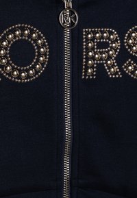 Marinblå zip-up hoodie med metalliska nitar, stora bokstäver kantade med nitar och en framträdande dragkedja med ett märkesdrag.