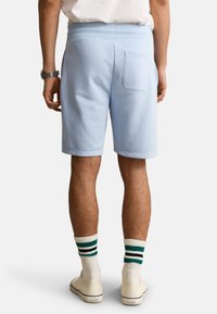 Homme portant un short bleu clair avec une poche arrière, des chaussettes blanches à rayures vertes et noires, des baskets blanches et une montre-bracelet.