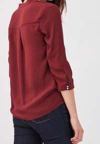 Bourgognebluse med trekvartlange ærmer, knapdetaljer på manchetterne, blødt stof, inkluderer en detalje med søm bagpå, kombineret med mørke jeans.