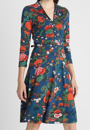 Kobieta w niebieskiej sukience do kolan z czerwonymi, zielonymi i białymi kwiatowymi wzorami, zapinana z przodu, z rękawami do łokcia i paskiem w talii.