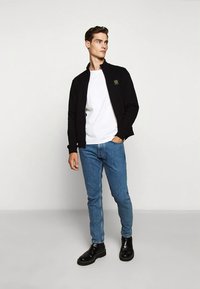 Casaco preto com fecho éclair por cima de uma t-shirt branca, combinado com jeans azuis e botins pretos até ao tornozelo. O casaco tem um pequeno emblema no peito.