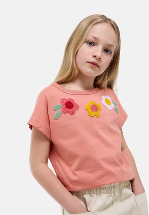 Jeune fille aux longs cheveux blonds portant un t-shirt rose à manches courtes avec trois motifs de fleurs texturées et un pantalon beige, les mains dans les poches.