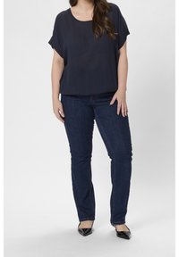 Donkerblauwe blouse met korte mouwen en een ontspannen pasvorm, gecombineerd met strakke donkere jeans en zwarte puntige hakken. Glad textuur overal.