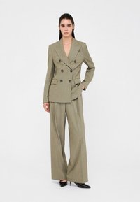 Blazer beige doppiopetto con grandi bottoni neri, abbinato a pantaloni a gamba larga nello stesso tessuto. Tacchi appuntiti neri ad alto contrasto.