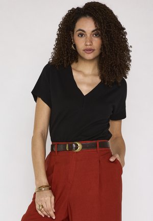 Donna con i capelli ricci che indossa un top nero con scollo a V, pantaloni a vita alta color ruggine con cintura marrone, gioielli d'oro, in piedi con una mano in tasca.