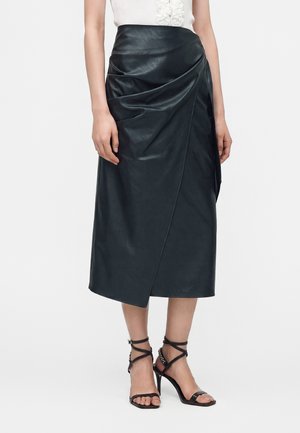 Vrouw draagt een zwarte midi rok van nepleer met geplooide details, gecombineerd met zwarte sandalen met hoge hakken en bandjes.