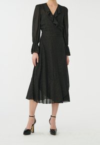 Schwarzes Wickelkleid mit Rüschendetails, langen Ärmeln und ausgestelltem Saum. Aus leichtem Stoff mit dezenter, strukturierter Musterung, kombiniert mit schwarzen High Heels.