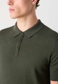 Mørkegrønn strikket polo skjorte med krage, korte ermer og en enkelt knapplukking ved halsen. Teksturert stoffdetalj er synlig.