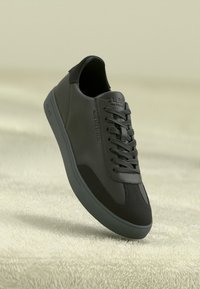 Black leather sneaker with textured fabric accents, rounded toe, and flat laces. Rubber sole is flat and matches the upper color.