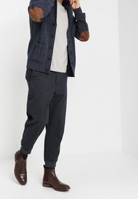 Cardigan blu navy a maglia cable-knit con toppe in pelle marrone sui gomiti, t-shirt grigia, pantaloni scuri e stivaletti alla caviglia in pelle marrone.