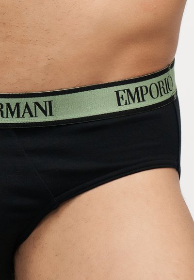 Buste masculin portant un slip noir avec une ceinture verte affichant le nom de la marque "EMPORIO ARMANI" en lettres noires.
