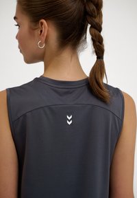 Junge Frau mit geflochtenem Haar, die von hinten zu sehen ist, trägt ein ärmelloses dunkelgraues Top mit einem kleinen weißen Doppelpfeil-Logo im oberen Rückenbereich.