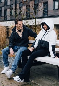 Two individuals sitting on a bench, wearing jackets—one black and white fleece with hood, the other dark with beige inner layer. Both in jeans and sneakers.