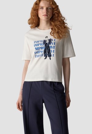 Woman with curly red hair wearing a white T-shirt with a blue "IMPREVE" print and a graphic of a woman in patterned clothing.