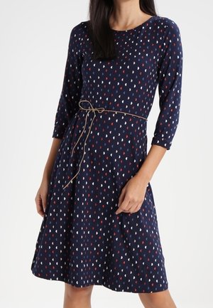 Kobieta ubrana w granatową sukienkę z białymi i czerwonymi wzorami w kształcie serc, z rękawami do łokcia i cienkim paskiem związanym w talii.