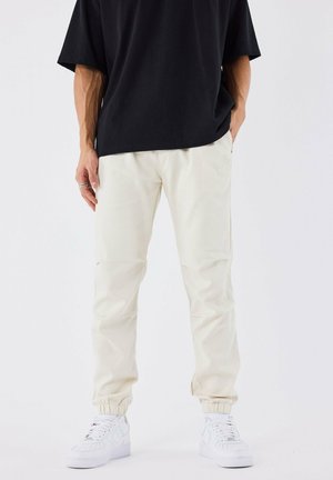 Pantalons crème à poignets élastiques avec une coupe décontractée, associés à un t-shirt noir surdimensionné et des sneakers blanches. Tissu lisse, détails minimaux.