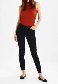 Černé slim-fit kalhoty s poutky na opasek, doplněné bezrukávovým červeným topem se žebrováním. Modelka stojí v černých balerínách a pózuje se zamotanýma rukama v kapsách.