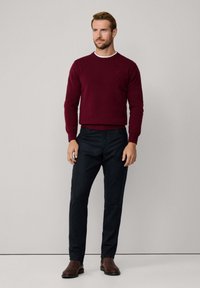 Un pull bourgogne avec un col rond, en tissu texturé, associé à un pantalon tailleur bleu marine et des bottes en cuir marron, se tenant devant un fond clair.
