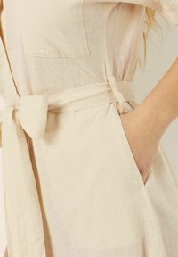Abito in lino beige chiaro con cintura da annodare, tasca laterale e vestibilità morbida. Presenta un design semplice e minimalista e una superficie morbida.