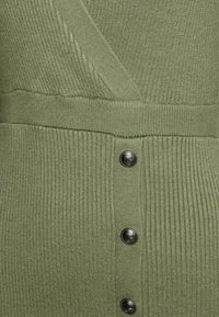 Gros plan sur un tissu en maille côtelée vert olive avec une couture diagonale et trois boutons métalliques ornés d'un motif circulaire.