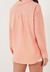 Chemise à manches longues rayée corail et blanche, dotée d'un empiècement dos, de poignets boutonnés et d'une coupe décontractée. Tissu doux avec des rayures visibles.